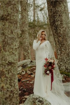 a woman in a white wedding dress standing next to trees and talking on a cell phone