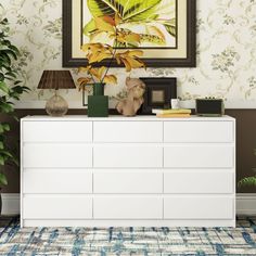 a white dresser sitting in front of a painting on the wall next to a potted plant