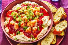 a bowl filled with food next to slices of bread on top of a purple table cloth