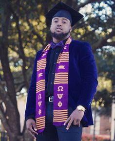 a man wearing a purple and yellow graduation stole standing in front of a tree with his hand on his hip