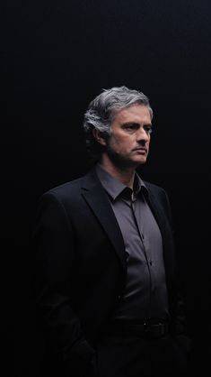 a man with grey hair wearing a black suit and tie standing in front of a dark background