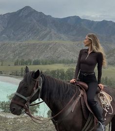 a woman riding on the back of a brown horse next to a lake and mountains