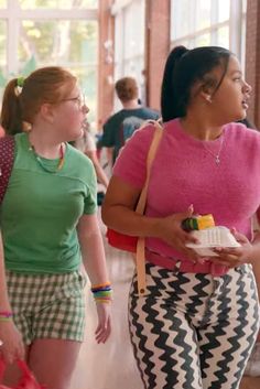 two women are walking down the hall with bags and food in their hands, one is carrying a cake