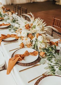 the table is set with white and orange flowers, gold place settings and napkins