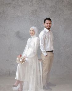 a man and woman standing next to each other in front of a gray wall wearing wedding attire