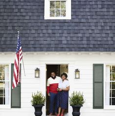 two people standing in front of a white house with an american flag on the porch