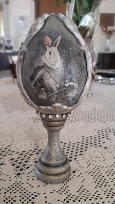 an old fashioned metal object with a rabbit on it's face and some flowers in the background