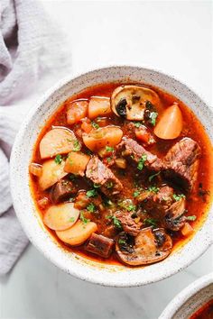 a bowl of stew with meat, potatoes and parsley in it on a white surface