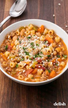 a white bowl filled with pasta and meat soup on top of a wooden table next to a spoon