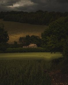 a house in the middle of a field with trees around it and dark clouds overhead