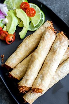 several burritos are on a black plate next to some vegetables and lime wedges