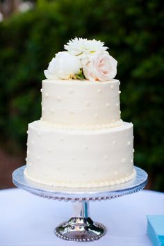 a wedding cake with white flowers on top is sitting on a blue tablecloth in front of some bushes