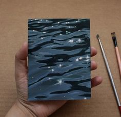 a hand holding a piece of paper with water and stars on it next to a paintbrush