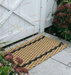 a door mat sitting on the ground next to some flowers and plants in front of a white fence