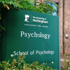 a green sign sitting in front of a brick building with trees and bushes around it