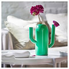 a green cactus vase with pink flowers on a table in front of a couch and pillows