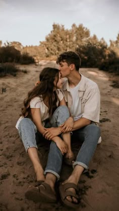 a man and woman sitting in the sand kissing each other with trees in the background