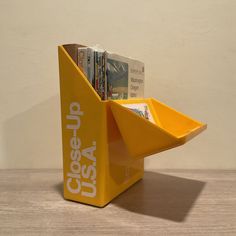 a yellow book holder with magazines in it sitting on a wooden table next to a white wall