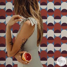 a woman holding a grapefruit in front of a wall with geometric designs on it