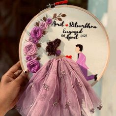 a hand holding up a purple dress with flowers on it and a embroidery hoop hanging from the wall