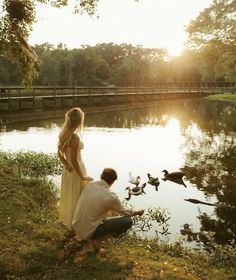 a man kneeling down next to a woman near a lake with ducks flying around it