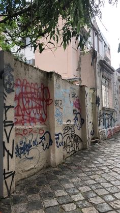 a wall covered in graffiti next to a brick sidewalk with trees and buildings behind it