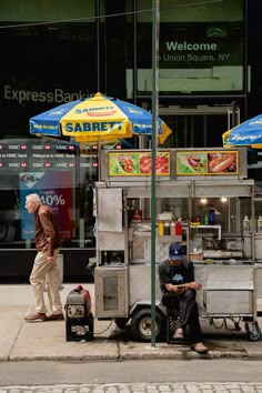 two men are sitting at a food cart on the sidewalk in front of a store