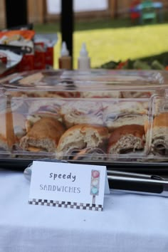 some sandwiches are in plastic containers on a white table cloth and there is a sign that says speedy sandwiches