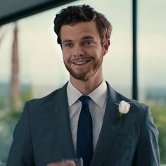 a man in a suit and tie holding a wine glass smiling at the camera while wearing a boutonniere