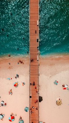 an aerial view of people on the beach and in the water near a pier with umbrellas