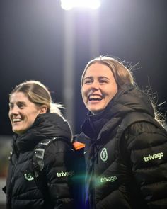 two women in black jackets standing next to each other on a soccer field at night