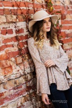 a beautiful woman leaning against a brick wall wearing a hat and dressy blouse with bell sleeves