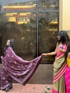 two women dressed in colorful sari are standing near a window and one woman is holding a shawl over her head