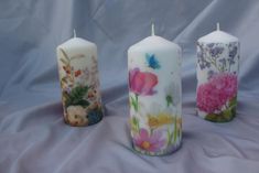 three candles with flowers painted on them are lined up against a blue background, one is white and the other is pink