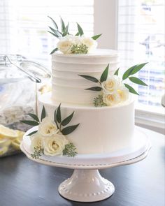 a wedding cake with white flowers and greenery on top sits on a table in front of a window