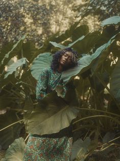 a woman standing in front of large leaves with her hands behind her head and looking up at the sky