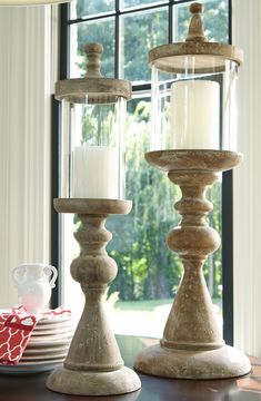 two candle holders on a table in front of a window with plates and cups next to them