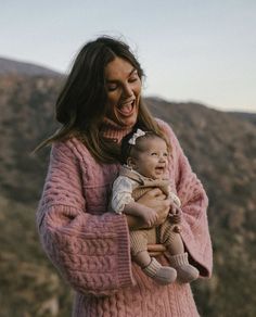 a woman holding a baby in her arms and laughing at the camera with mountains in the background