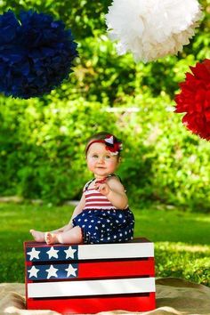 a baby sitting on top of a wooden box with red, white and blue pom poms