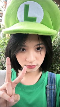 a young woman wearing a green hat making the peace sign with her hand while standing in front of bushes