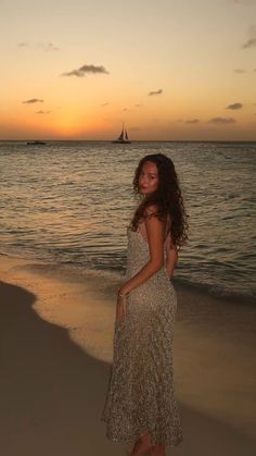 a woman standing on top of a sandy beach next to the ocean at sunset with a sailboat in the distance