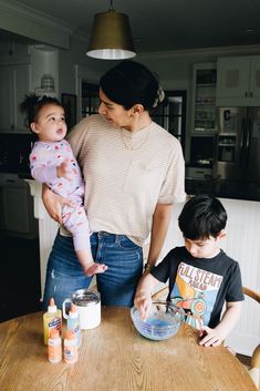 a woman holding a baby while standing next to two other children at a kitchen table