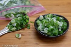 chopped green onions in a black bowl next to a spoon on a wooden table with plastic bags