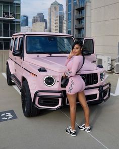 a woman standing next to a pink mercedes g - class in front of a cityscape