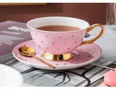 a pink and gold coffee cup on a saucer next to a book with a spoon in it