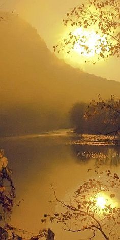 the sun shines brightly through the foggy sky over a lake with trees and mountains in the background