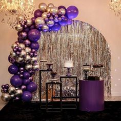 a purple and silver balloon arch with candles on the table in front of it, surrounded by balloons