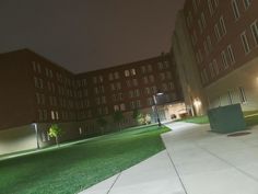 an empty sidewalk in front of a large building at night with lights shining on the grass