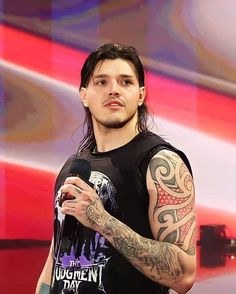 a man with long hair and tattoos holding a microphone in front of a tv screen