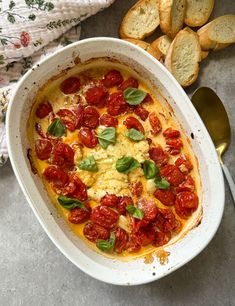 a casserole dish with tomatoes and cheese in it next to bread on the side